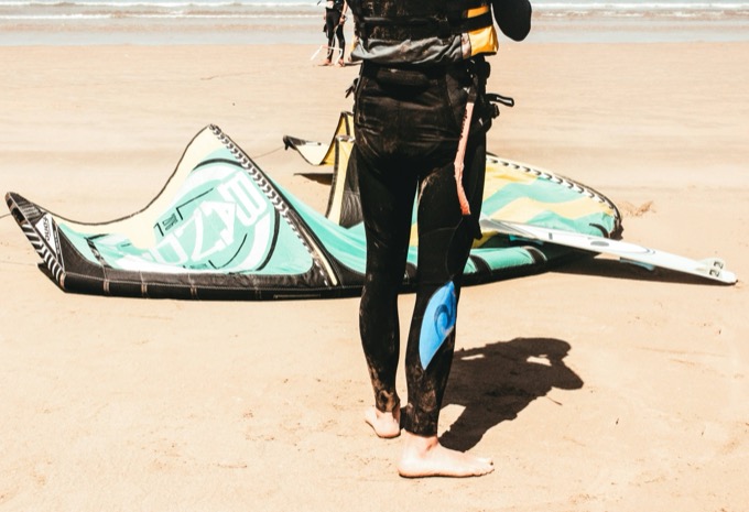 Kitesurfer in a black wetsuit standing beside kite equipment on the beach